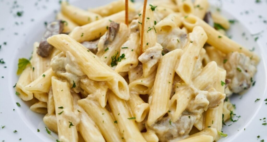 Closeup shot of penne pasta coated in a creamy Alfredo sauce. The dish includes pieces of mushrooms and is garnished with finely chopped parsley, adding a touch of color. The creamy sauce clings to each piece of pasta, highlighting its rich and indulgent texture