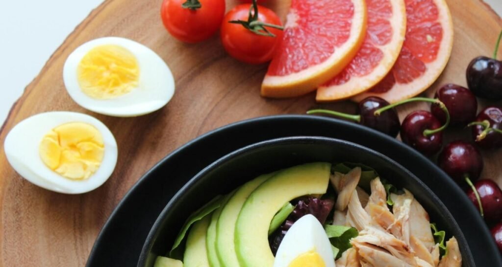 Healthy meal featuring sliced avocado (a source of healthy fats), boiled eggs, cherry tomatoes, grapefruit slices, cherries, and chicken in a black bowl, emphasizing a balanced diet for weight loss.