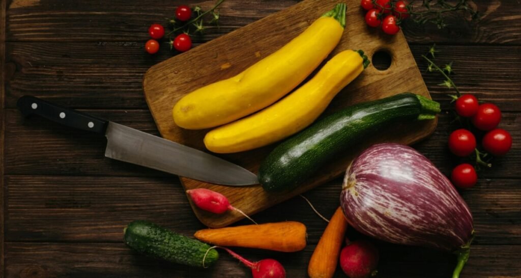 Fresh yellow squash, zucchini, radishes, and other vegetables on a wooden cutting board with a knife, surrounded by cherry tomatoes.